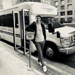 A picture of Nathan Segura standing in front of a Santa Barbara Airbus and leaning on a sign attached to the sidewalk for "Artists Only"